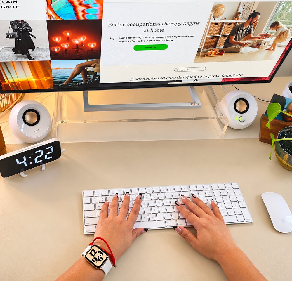 Jennifer’s hands typing at her desk, with the Kinspire website on a large monitor, a clock reading 4:22 and a plant nearby