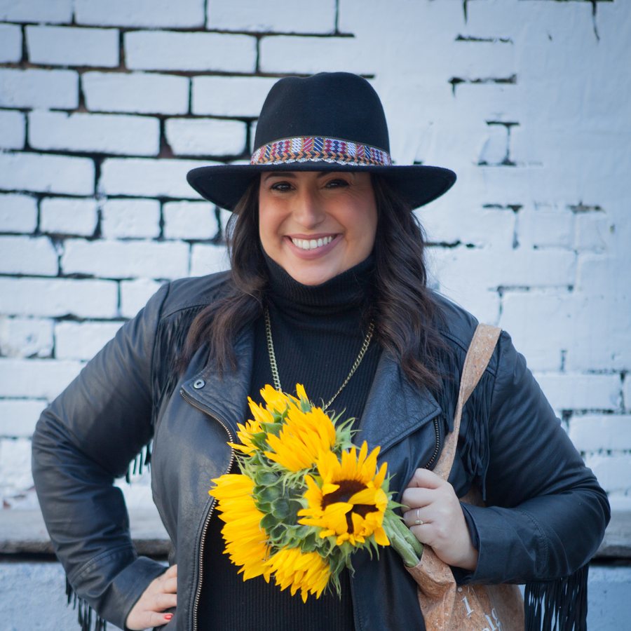 Jennifer smiling in a black hat and fringed leather jacket, holding a bunch of sunflowers against a white brick wall